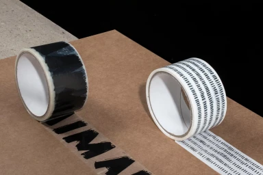Two rolls of patterned adhesive tape unrolling across a brown cardboard surface in a studio setting.