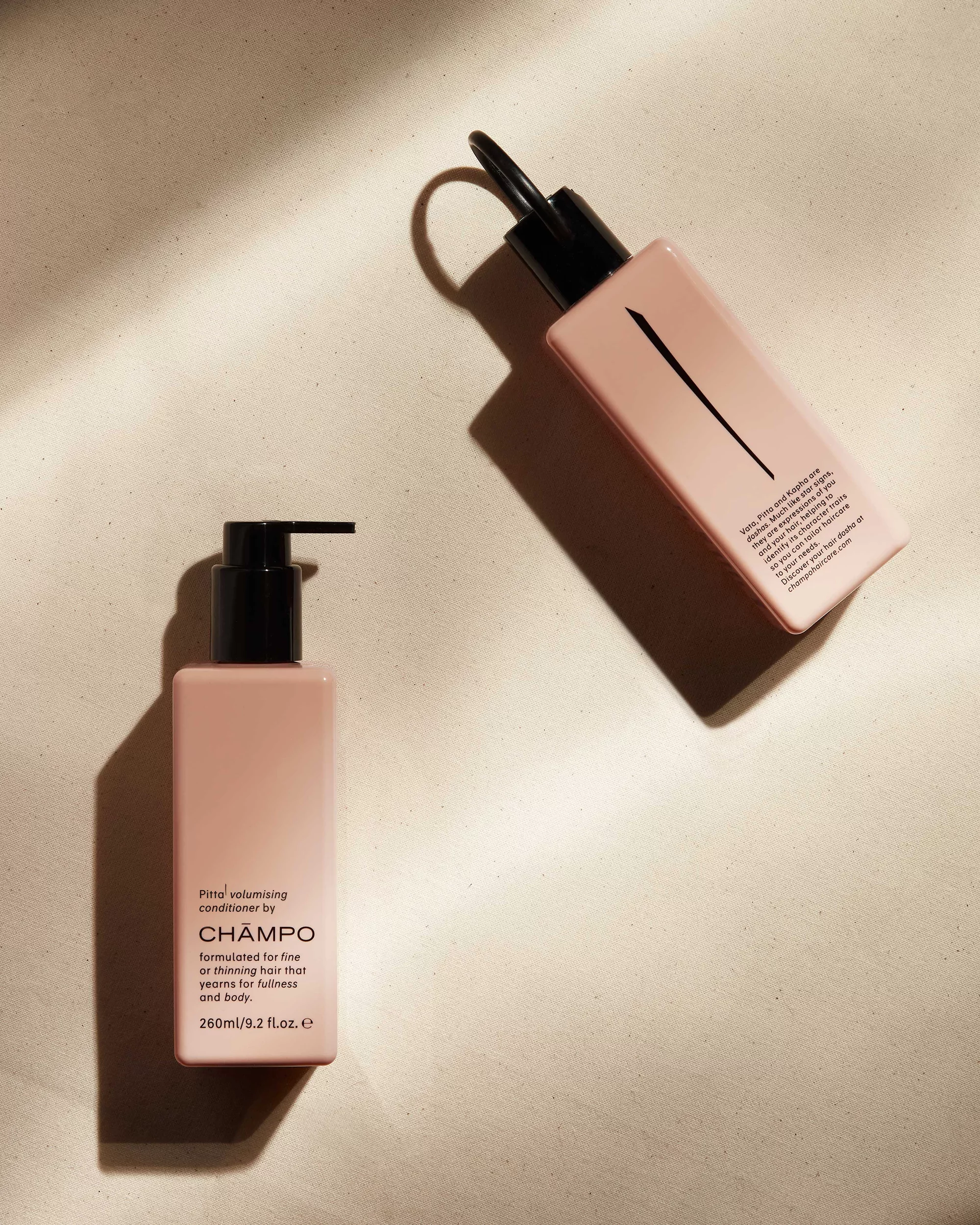 Still life photo of two light pink rectangular product bottles with black caps, one with a pump, on a cream colored surface with shadows.