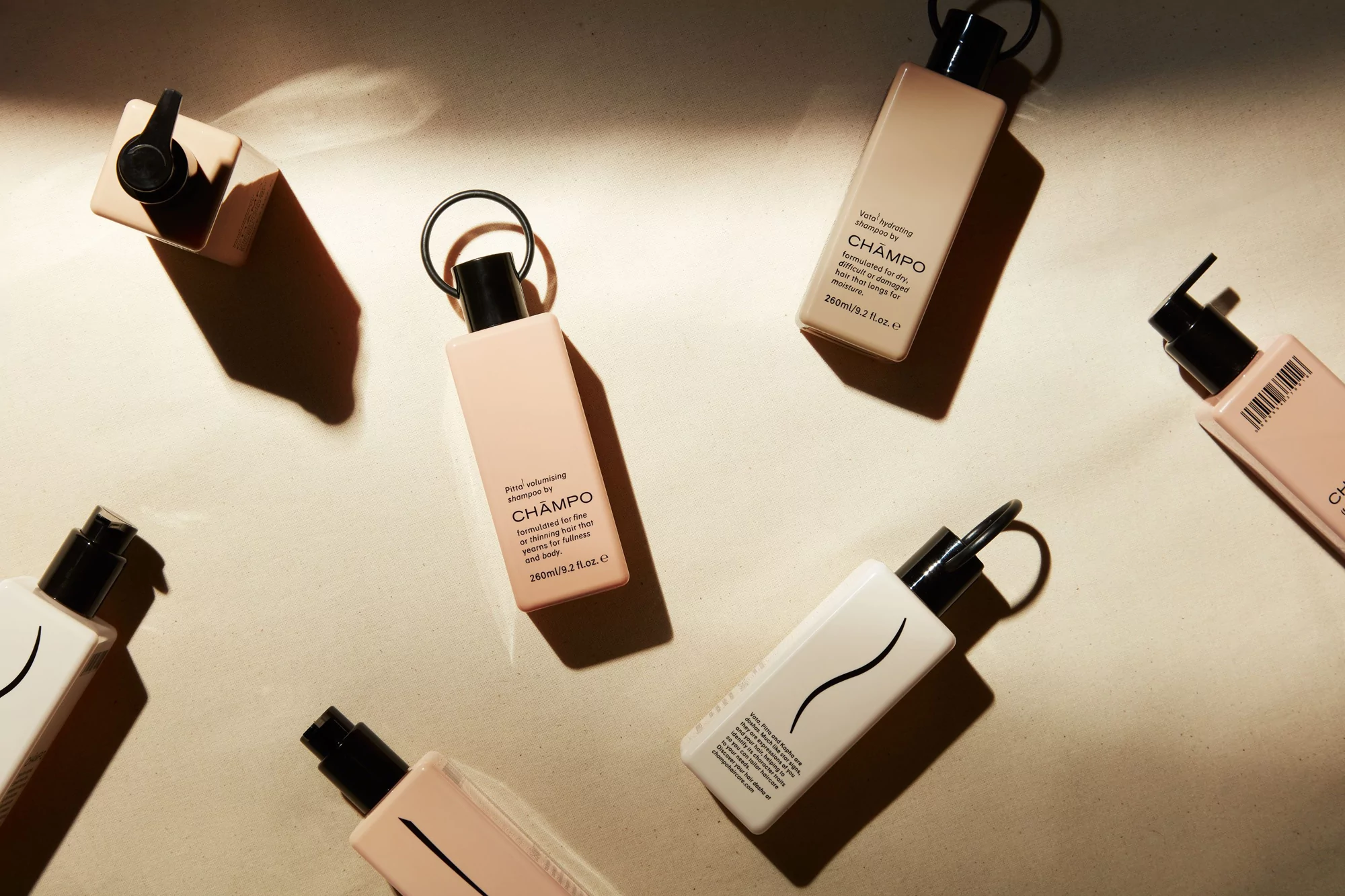 Overhead shot of several peach and white Chāmpo shampoo bottles with black caps, arranged on a beige surface with soft lighting.