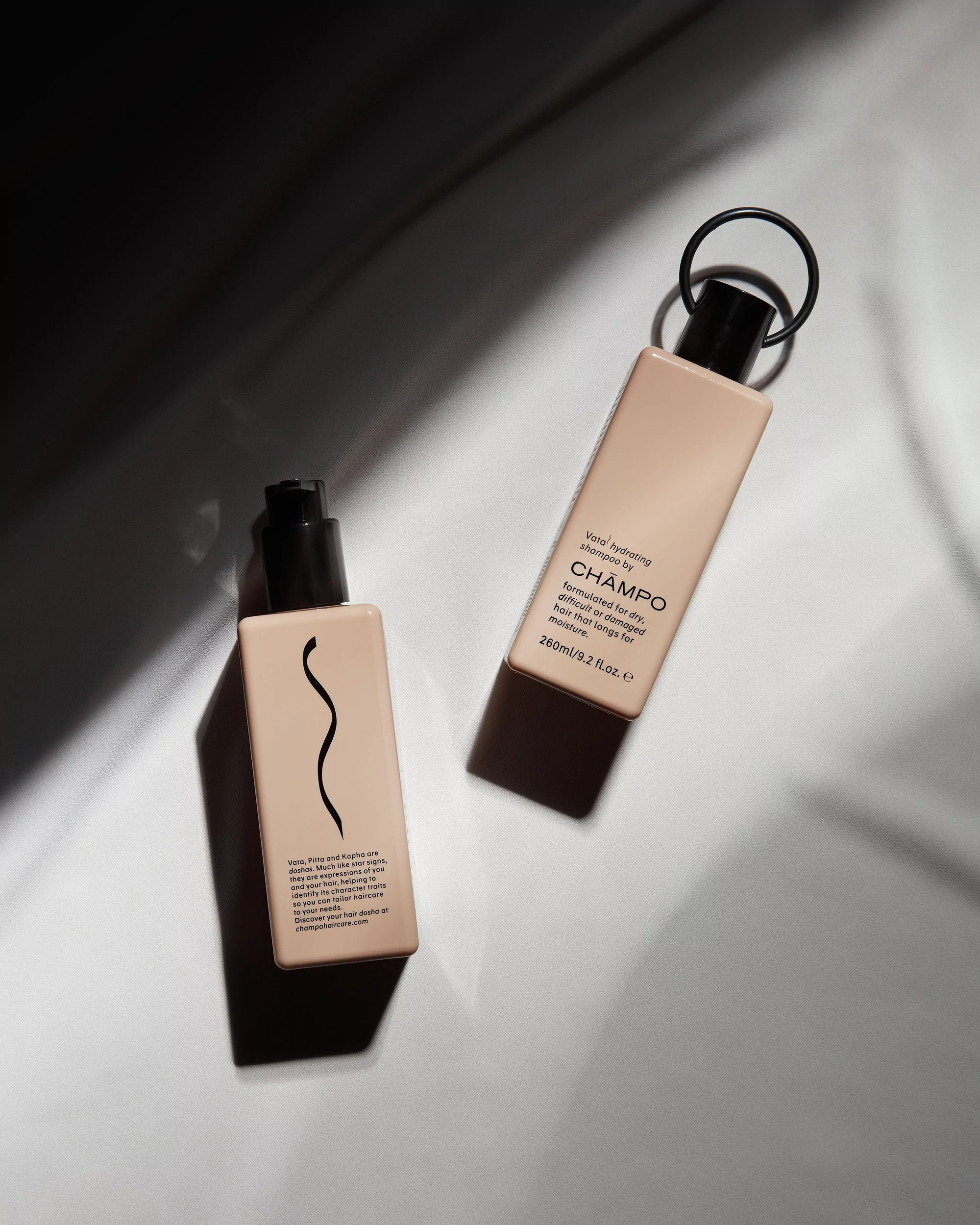 Two bottles of beige shampoo are displayed against a white background, with dark shadows creating an abstract design element in the scene.