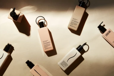 Top-down view of six Champo shampoo bottles arranged on a neutral stone surface with warm sunlight casting sharp shadows.