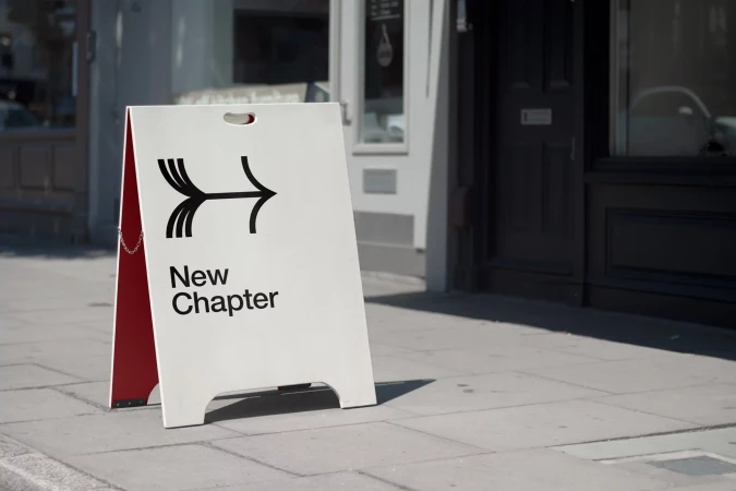 Outdoor shot of a sandwich board sign on a sidewalk with the text "New Chapter" and an abstract arrow graphic.