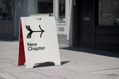 White sandwich board sign with black New Chapter branding on a concrete pavement outside a shop front.
