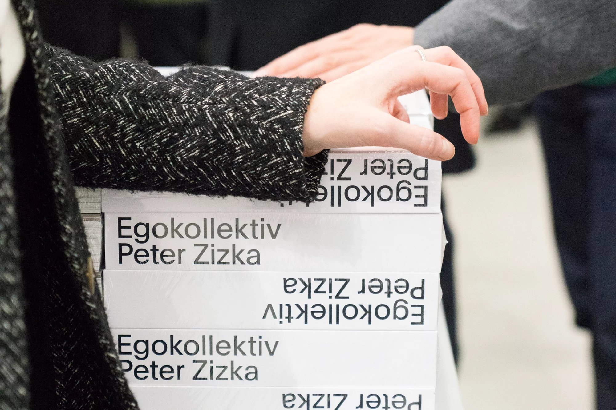 Close-up of a hand reaching for a stack of 'Egokollektiv Peter Zizka' books, with a focus on the books and hand interaction.