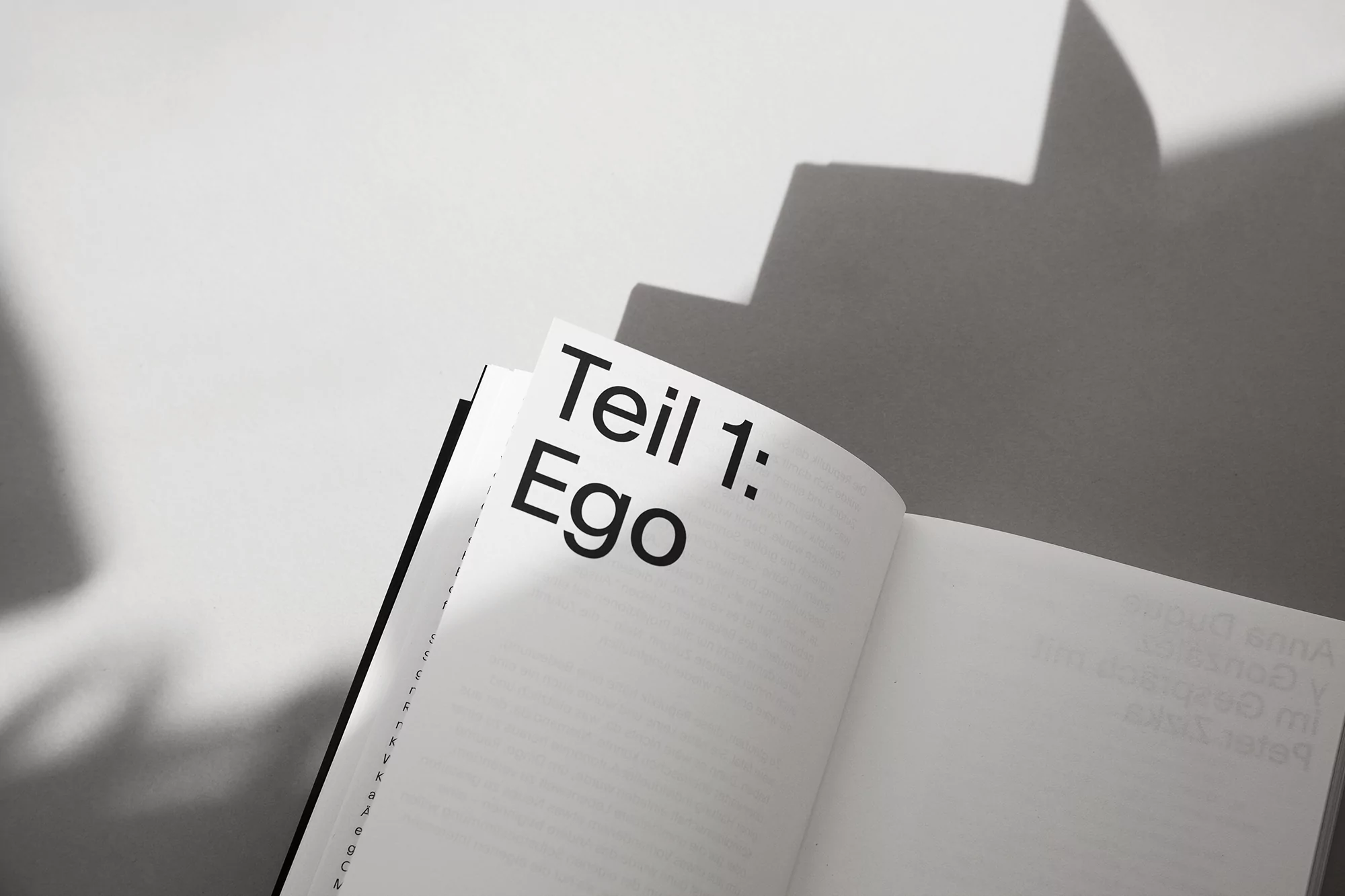Open book featuring the text 'Teil 1: Ego' in bold black letters, casting a sharp shadow on a white surface, minimalist.