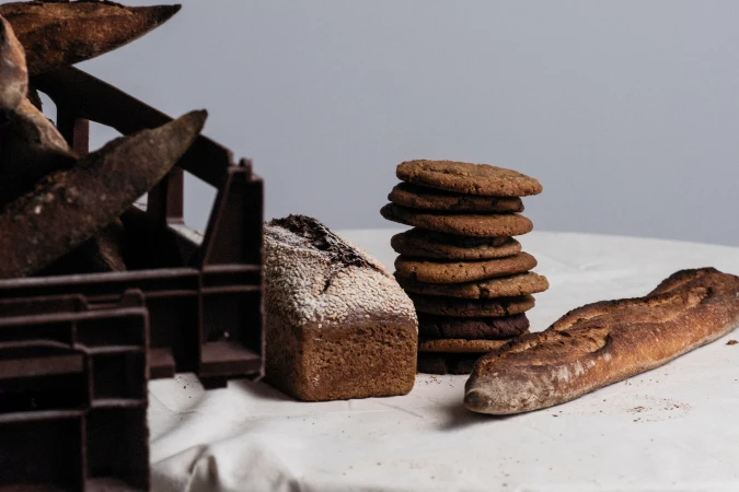 Artisan breads and cookies are displayed on a white tablecloth, with baguettes in a brown crate and stacked cookies near loaf.