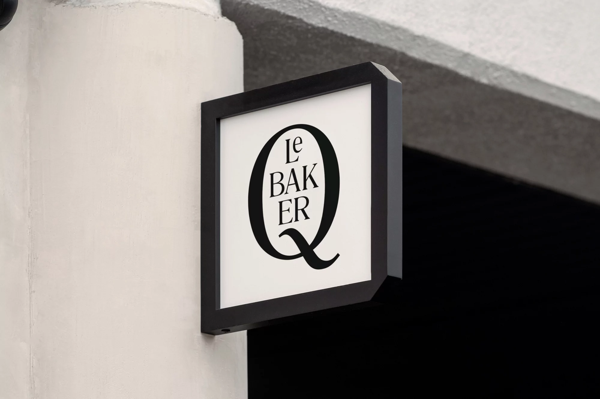 Close-up on a square business sign hanging on a cream-colored exterior wall, displaying the text 'Le Baker Q' in black font.