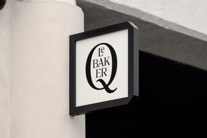 Close-up on a square business sign hanging on a cream-colored exterior wall, displaying the text 'Le Baker Q' in black font.