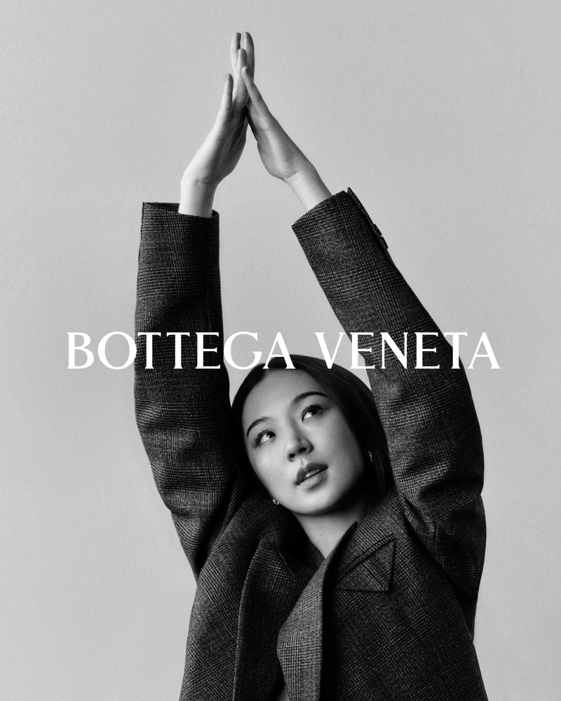 Black and white portrait of a woman in a blazer with her hands raised above her head, posing in front of a solid light-grey backdrop.