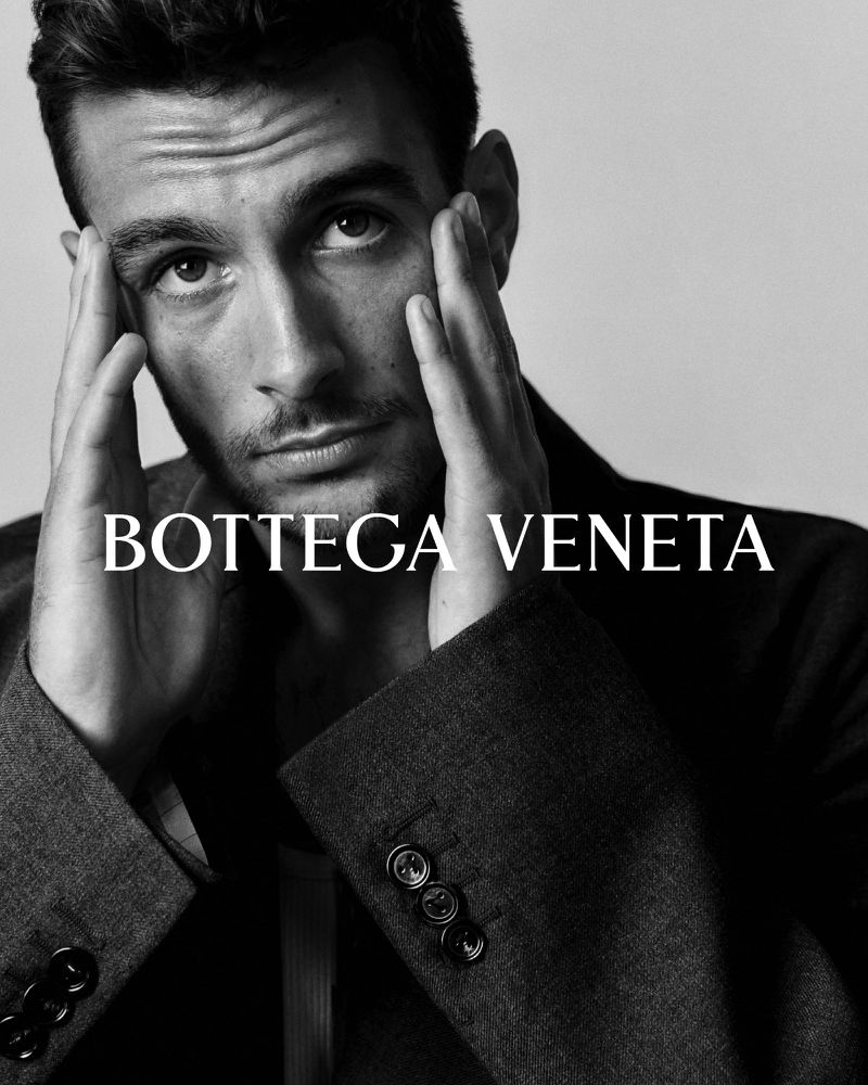 Portrait of a man in a dark blazer with hands near his face, posing in front of a light gray background, Bottega Veneta text overlay.