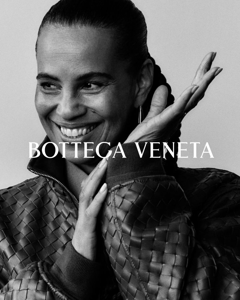 A smiling woman wearing a woven leather jacket poses for a black and white close-up studio portrait, showcasing fashion and beauty.