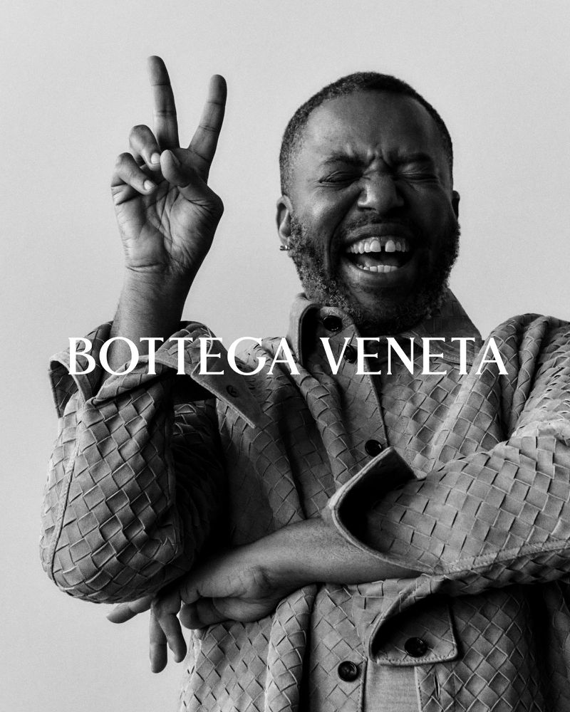 Black and white portrait of an adult male smiling and making a peace sign while wearing a woven coat from Bottega Veneta.