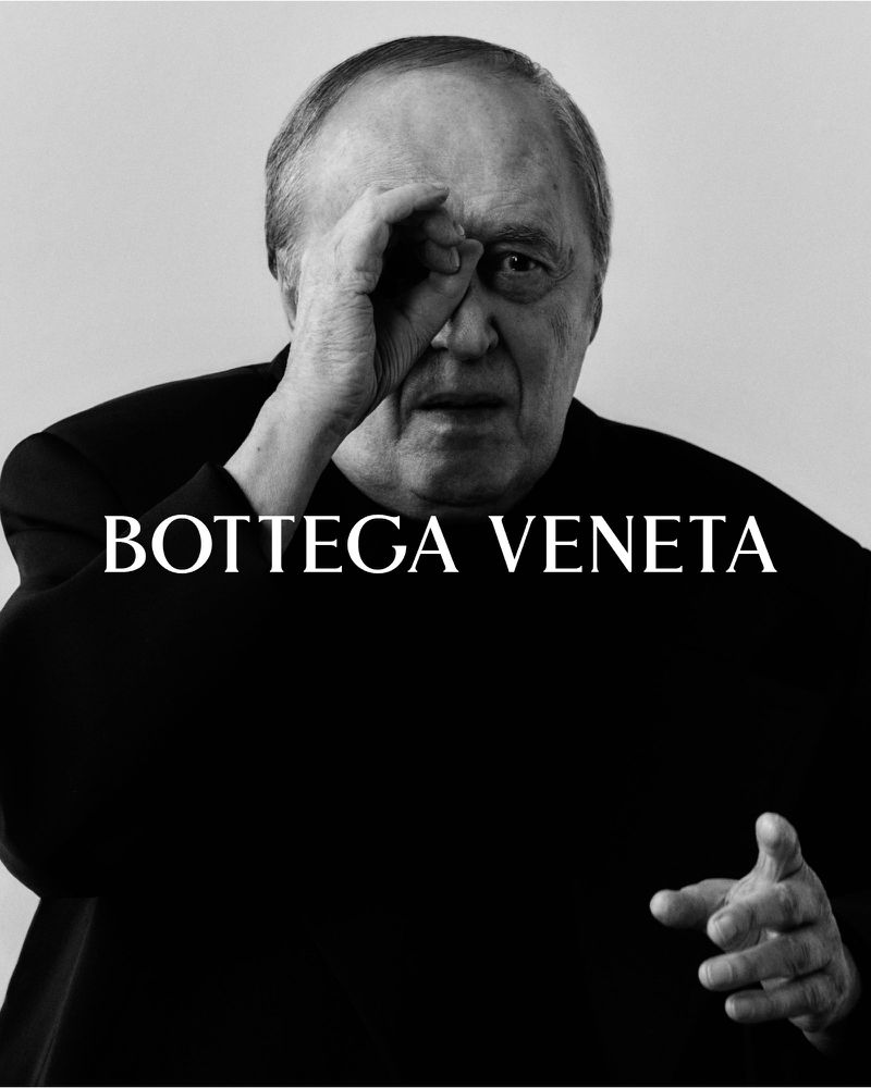 Black and white portrait of older adult male gesturing with hand, wearing black top in front of a plain background with brand text.