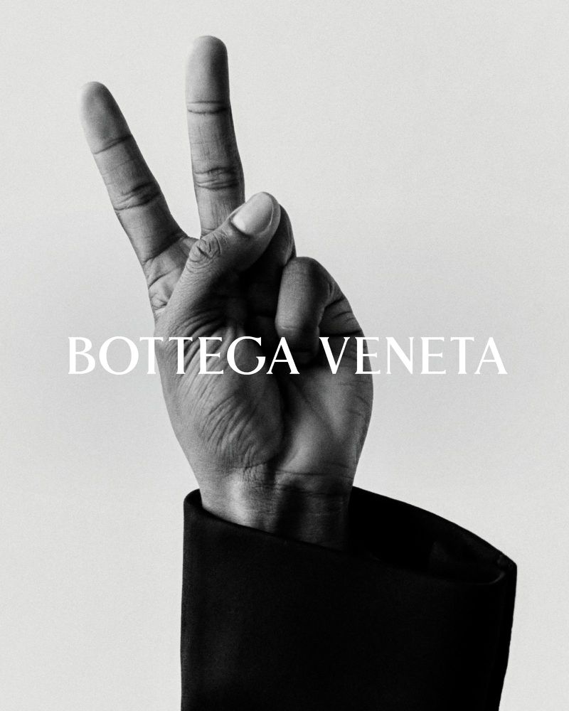 Bottega Veneta ad featuring a hand in a black jacket sleeve making a peace sign against a grey background with the brand name displayed.