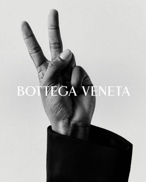 Bottega Veneta ad featuring a hand in a black jacket sleeve making a peace sign against a grey background with the brand name displayed.