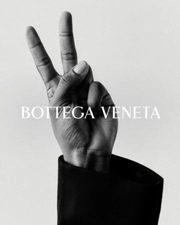 Bottega Veneta ad featuring a hand in a black jacket sleeve making a peace sign against a grey background with the brand name displayed.