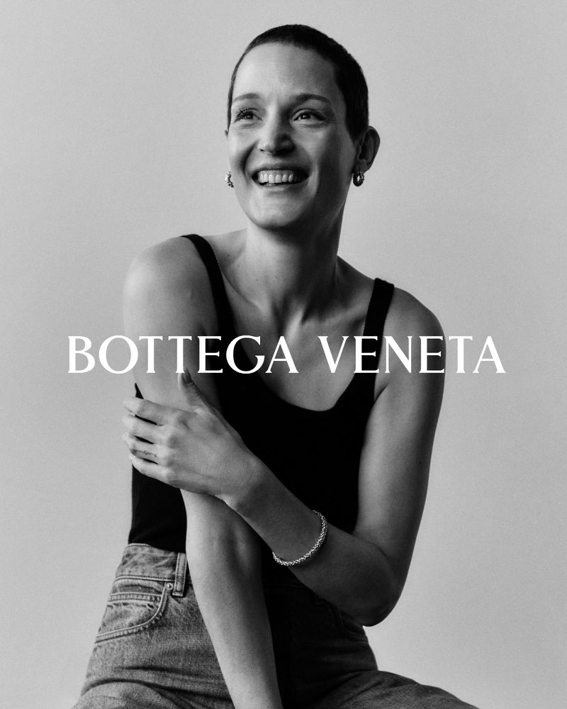 Black and white portrait of a smiling adult woman with short hair, wearing a tank top, jeans and a bracelet. "BOTTEGA VENETA" text overlay.