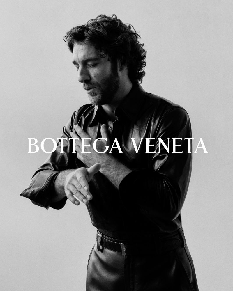 Black and white portrait of a man with curly hair wearing a black leather shirt, posing against a neutral backdrop in a photo studio.
