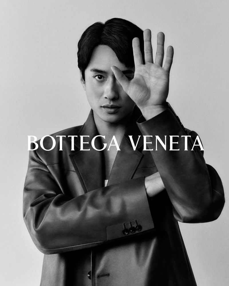Monochromatic portrait of an Asian male model in a Bottega Veneta jacket, shielding one eye with his hand, set against a neutral backdrop.