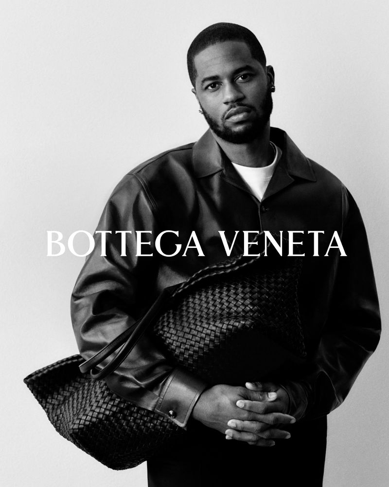 Black and white fashion portrait of a stylish Black man holding a woven leather Bottega Veneta bag against a neutral background.