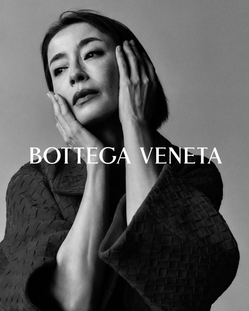 Close-up portrait of an Asian woman with hands on her face, wearing a textured jacket, in a black and white studio shot with brand logo.