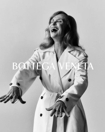 Black and white portrait of an adult woman laughing, wearing a white trench coat, set against a neutral background, featuring Bottega Veneta text.