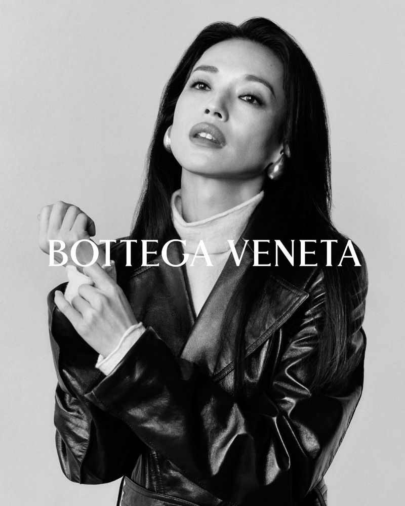 Headshot of an adult woman with dark hair wearing a black leather coat, white turtleneck, and earrings, posing in a photo studio.
