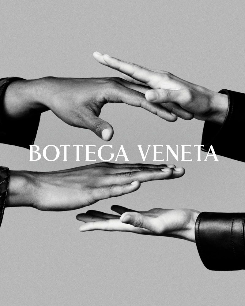 Three hands of different skin tones reach out, conveying unity and connection in a black-and-white studio shot, featuring Bottega Veneta logo.