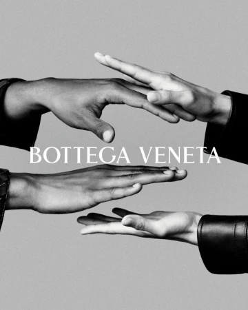 Three hands of different skin tones reach out, conveying unity and connection in a black-and-white studio shot, featuring Bottega Veneta logo.
