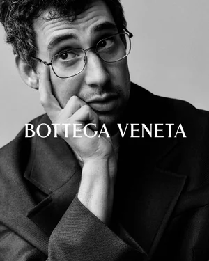 Portrait of a man wearing eyeglasses resting his hand on his cheek against a neutral background with Bottega Veneta text.