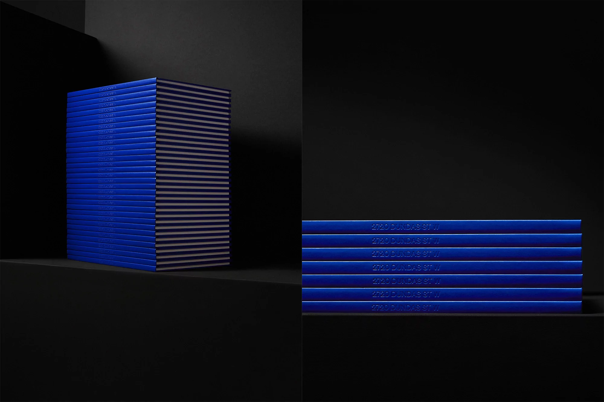 Stack of glossy cobalt blue books with 2720 DUNDAS ST. W printed, positioned on a black surface against a dark, minimalistic background.