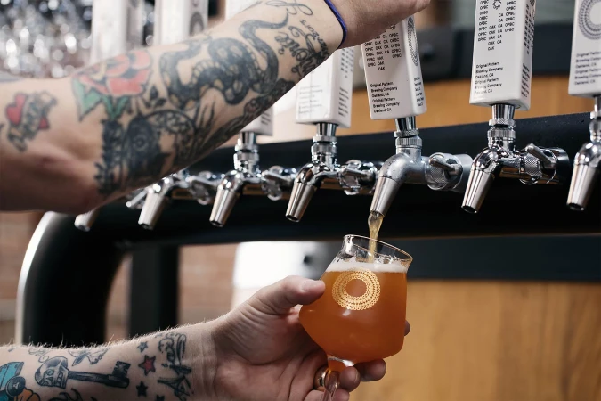 Craft beer pouring from a tap into a branded glass, held by a person with arm tattoos, in a brewery or bar setting.