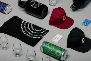 Flat lay brand identity items including branded t-shirts, caps, beer cans, bottles, and glasses arranged on grey background.