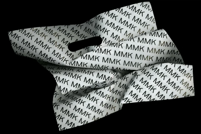 Stock image of a crumpled white shopping bag with an all-over print of repeating MMK letters in black, set against a stark black backdrop.