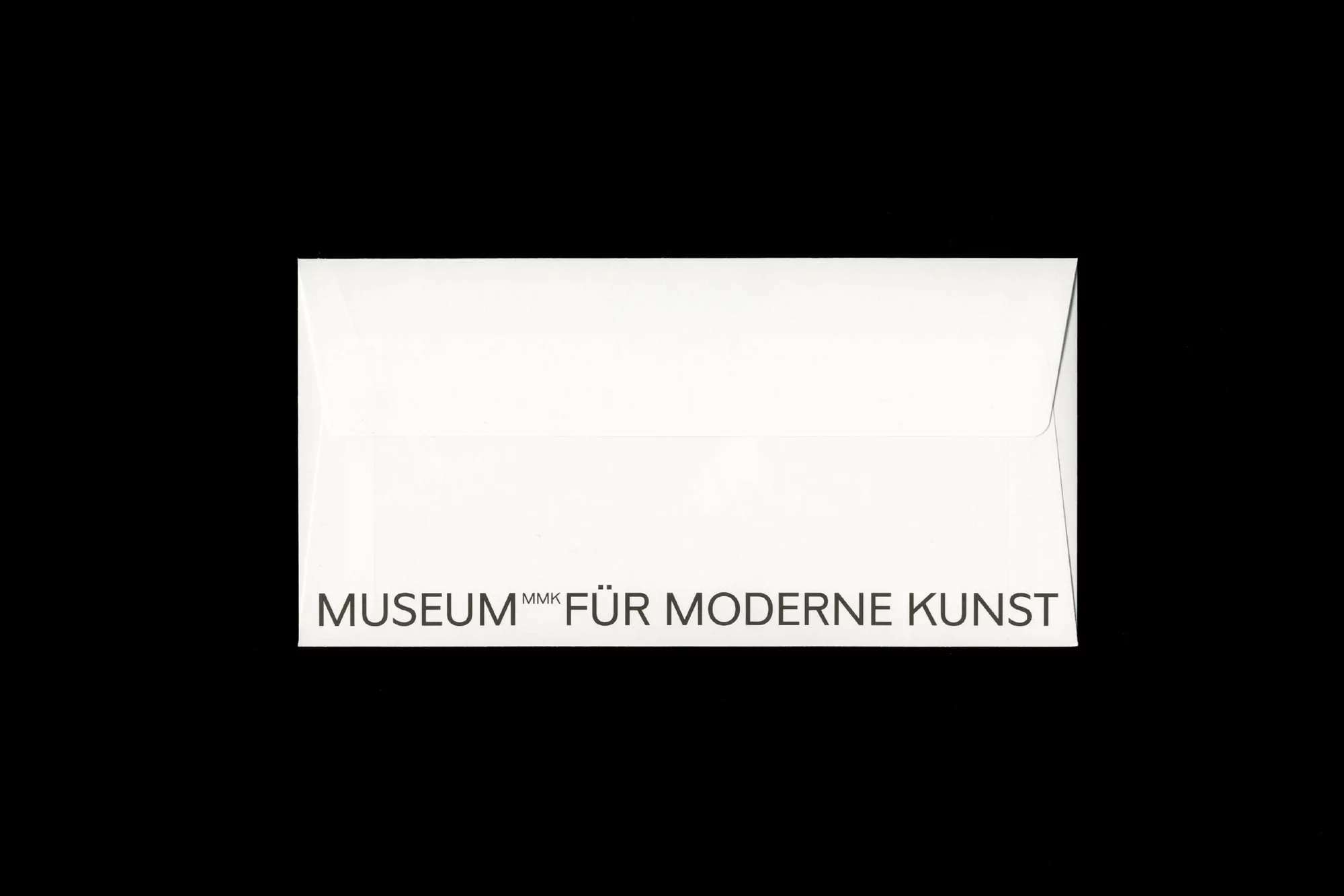 An envelope, with the words MUSEUM MMK FÜR MODERNE KUNST written in black on it, is set against a black background in a centered studio shot.