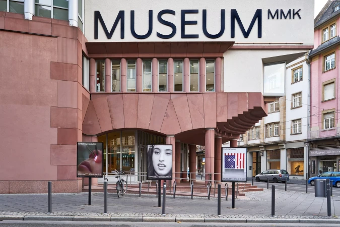 Facade of the MMK Museum in Frankfurt, Germany, with posters displayed on the sidewalk in front of the entrance.