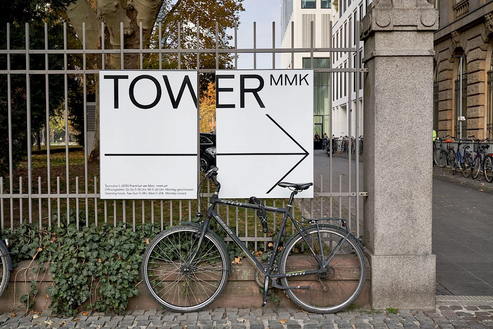 A bicycle parked in front of a sign for TOWER MMK contemporary art gallery in Frankfurt, Germany with an urban street scene backdrop.