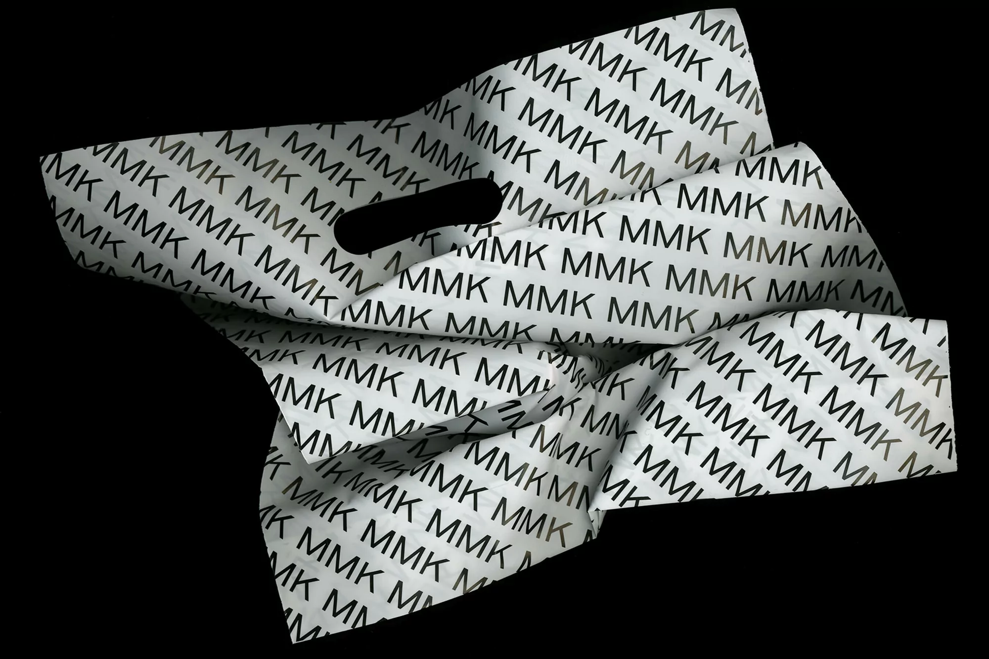 Crumpled white plastic carrier bag printed with repeating black MMK logotype pattern against a black background