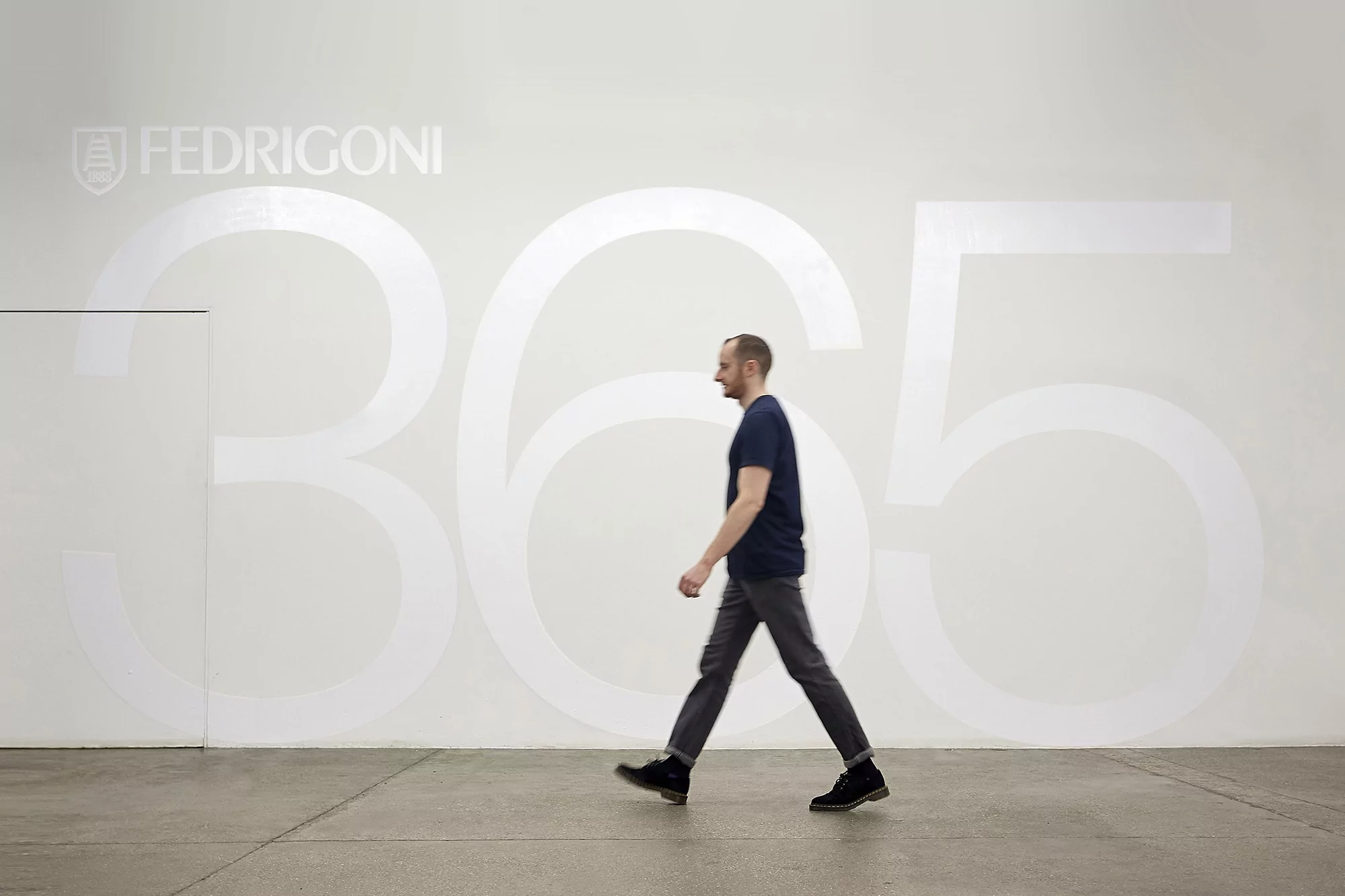 Adult male walks to the left in front of a white wall with 'FEDRIGONI' and '365' in translucent font on a grey concrete floor.