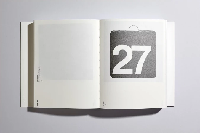 Open book on a white surface displays a calendar page with the number 27 in a bold, minimalist design, set against a gray square.