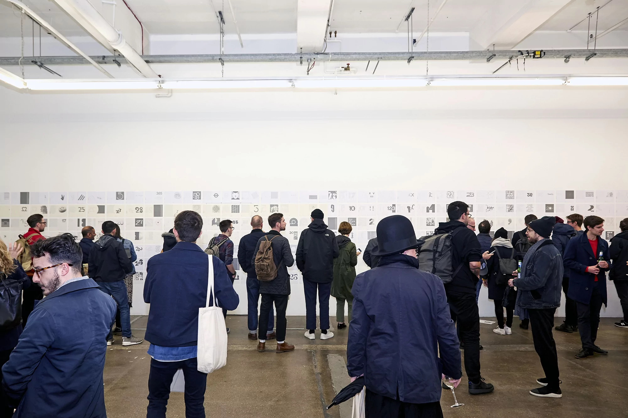 Large group of people at an indoor art exhibition looking at drawings hung on a white wall, neutral lighting, and concrete floor.