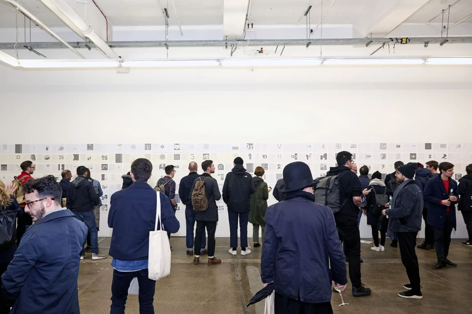 Large group of people at an indoor art exhibition looking at drawings hung on a white wall, neutral lighting, and concrete floor.