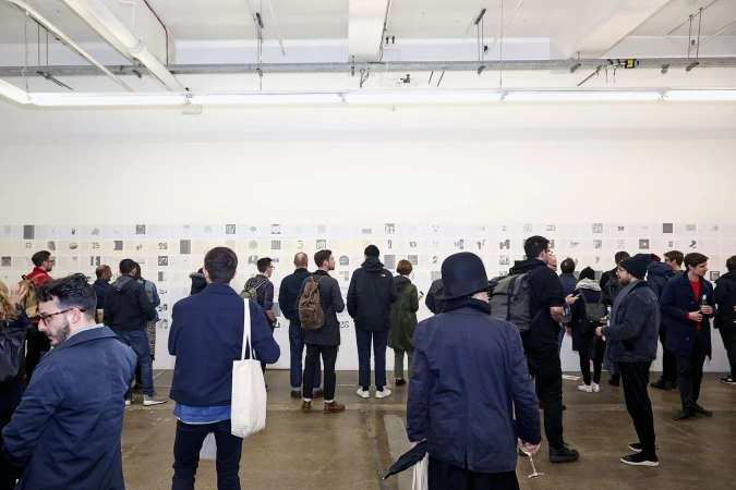 Large group of people at an indoor art exhibition looking at drawings hung on a white wall, neutral lighting, and concrete floor.