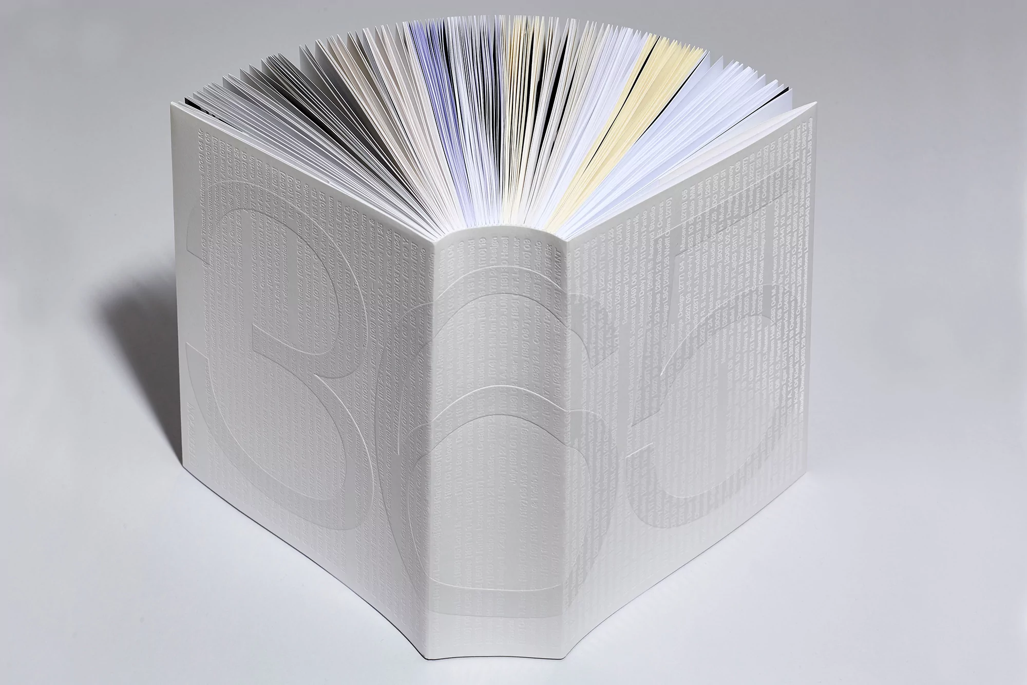 Close-up of a book with embossed cover, open to the middle, displaying pages with white, blue, and yellow edges on a white surface.