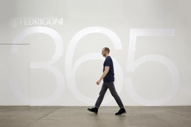 A man walking across a grey concrete floor in front of a white wall featuring the Fedrigoni logo and the number 365.