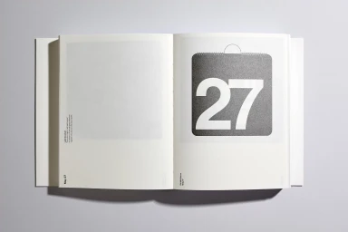 An open calendar book laying flat on a neutral grey surface displaying the number 27 in bold white text.