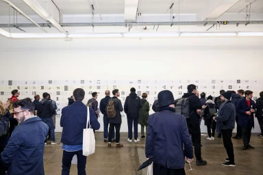 A crowd of people in an industrial exhibition space viewing graphic identity designs mounted on a white wall.