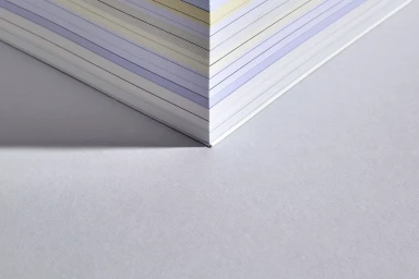 Close-up corner view of stacked pastel-colored paper sheets casting a sharp shadow on a grey surface.