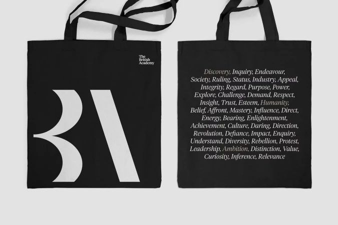 Two black tote bags featuring white text and logos are displayed on a white background. One bag has the letters BA in large font.