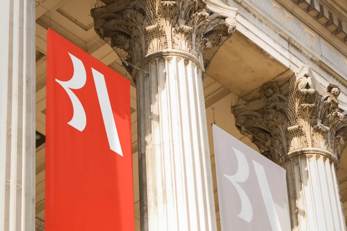 Architectural photograph of banners hanging between stone pillars, featuring a building exterior with ornate architectural details.